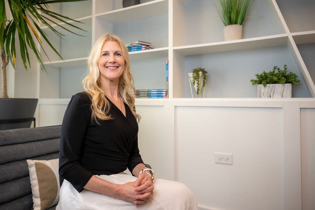 Darcie Beyer seated in the treatment room at All Is Well Austin, beside a shelf of plants and books.