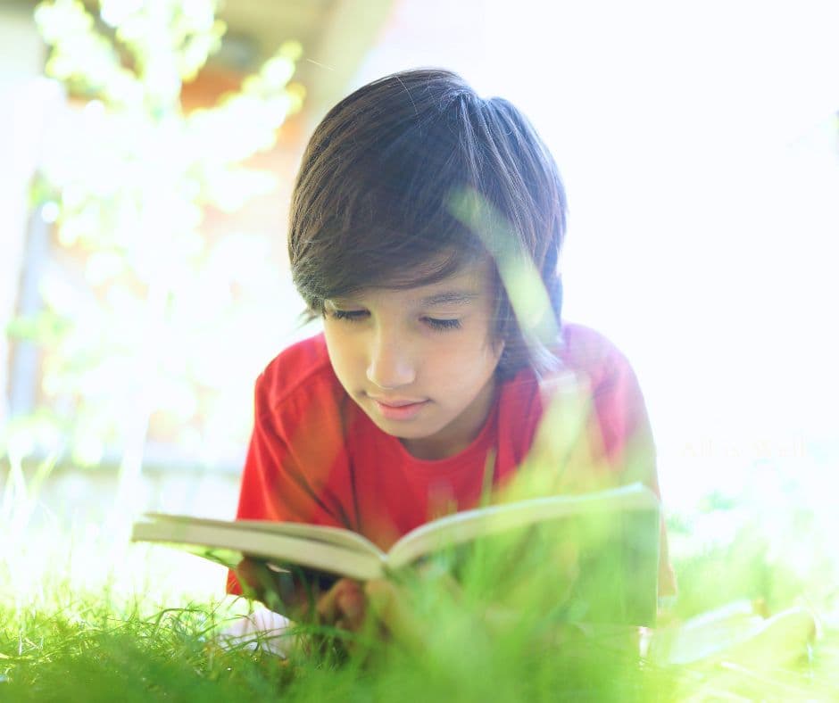 A boy lying in the grass, absorbed in the book he is reading.