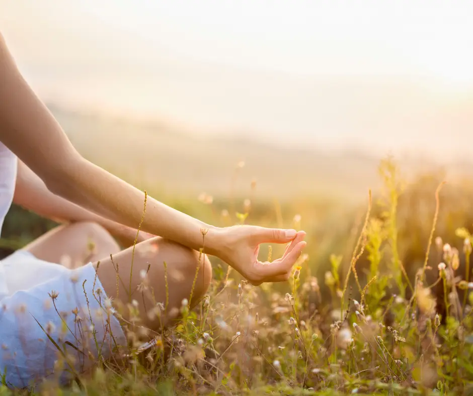 A person meditating cross-legged in a sunlit meadow at golden hour, one hand resting on the knee.