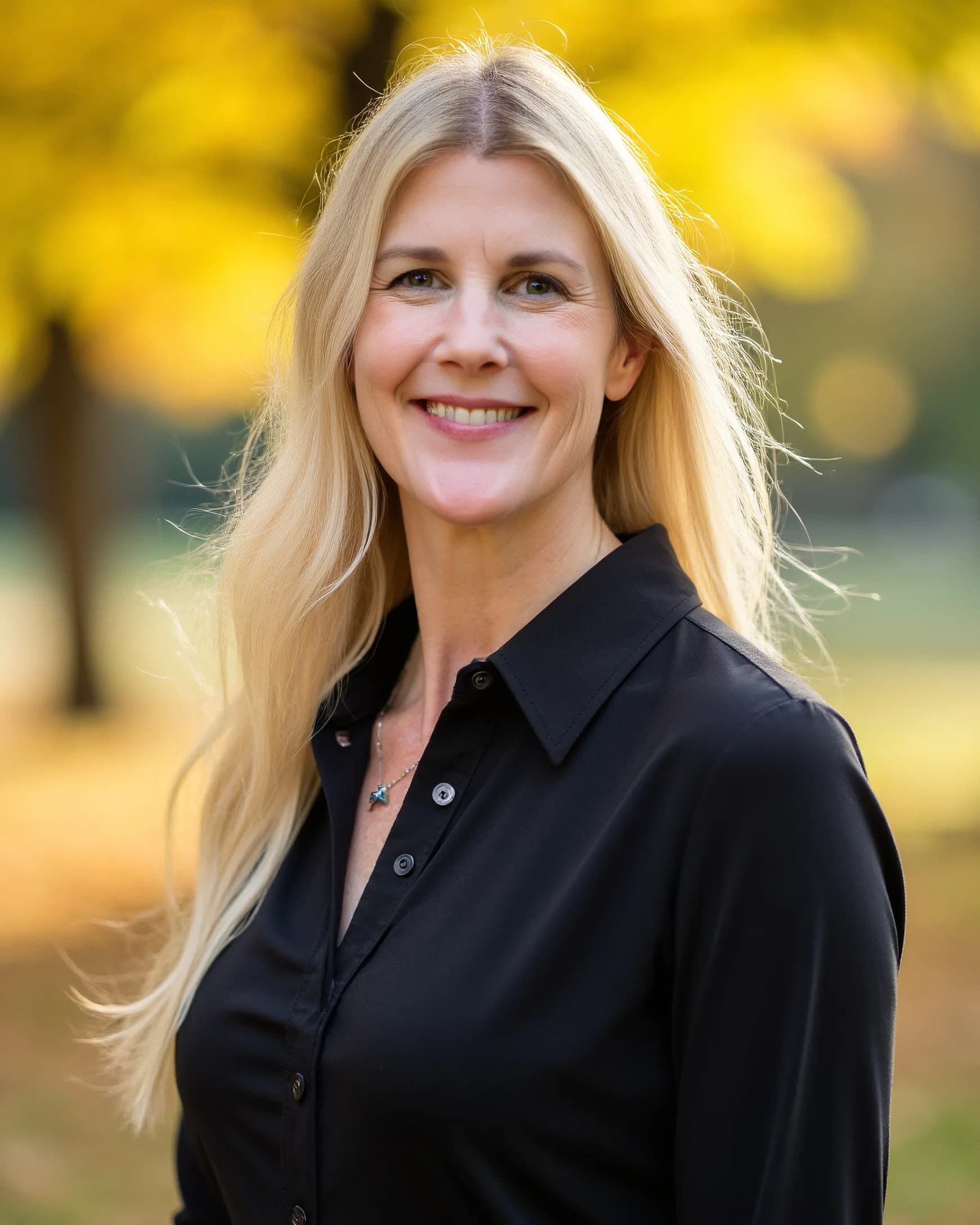 Darcie Beyer, L.Ac., founder of All Is Well Austin, standing outdoors in a black shirt, smiling, with autumn leaves soft behind her.