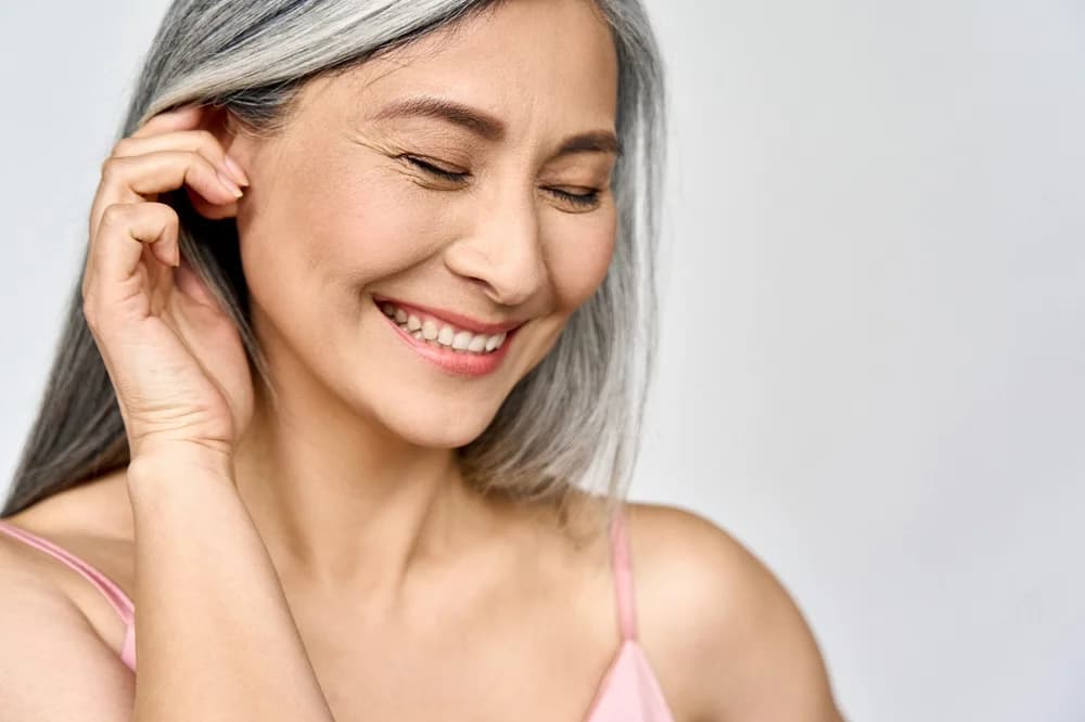 A smiling woman with long silver hair tucking it behind her ear, her skin relaxed and clear.